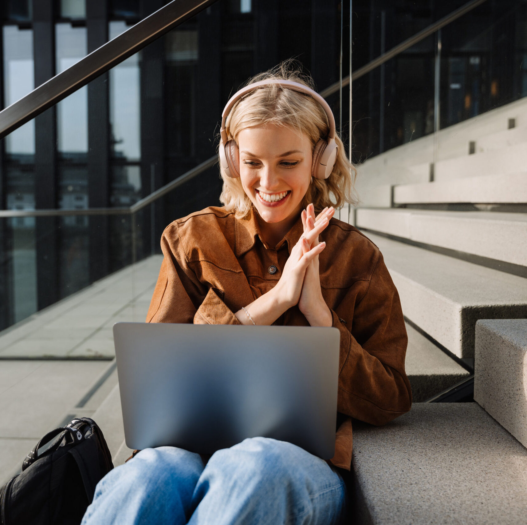 Young smiling blonde woman in wireless headphones making video call via laptop and gesturing while sitting on stairs outdoors